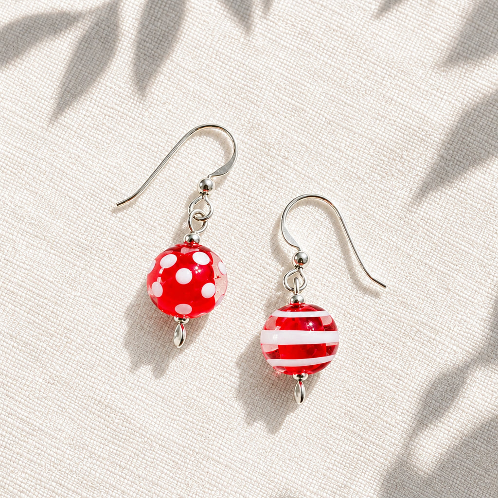 A pair of dangling earrings with silver fishhook wires and round glass beads. One bead is bright red with white polka dots, and the other is red and white striped.