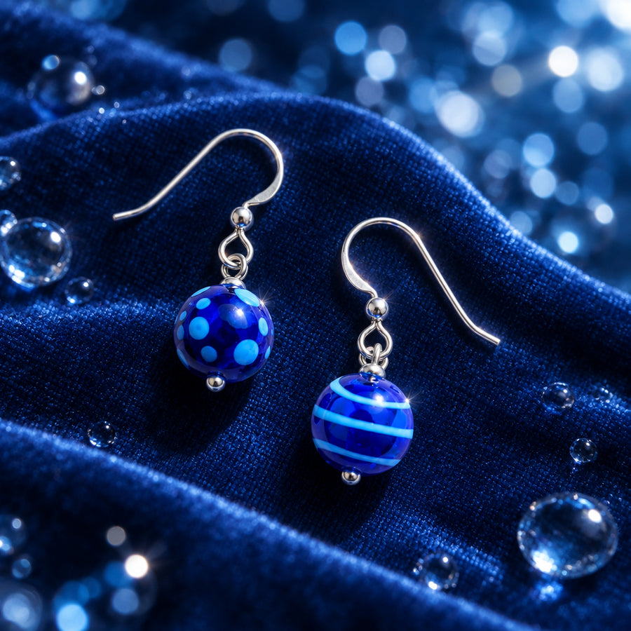 A pair of dangling earrings with polished silver-tone fishhook wires and glossy blue glass beads. One bead is deep cobalt blue with lighter blue polka dots, and the other is dark blue with horizontal light blue stripes.
