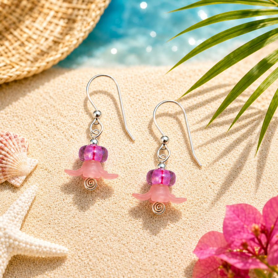 A pair of dangling earrings displayed on sandy beach-style props, with a woven hat edge, starfish, shells, palm leaves, and bright pink flowers around them. The earrings have silver fishhook wires, small silver beads, a translucent pink bead, a pink flower-shaped charm, and a small spiral gold-toned detail at the bottom.