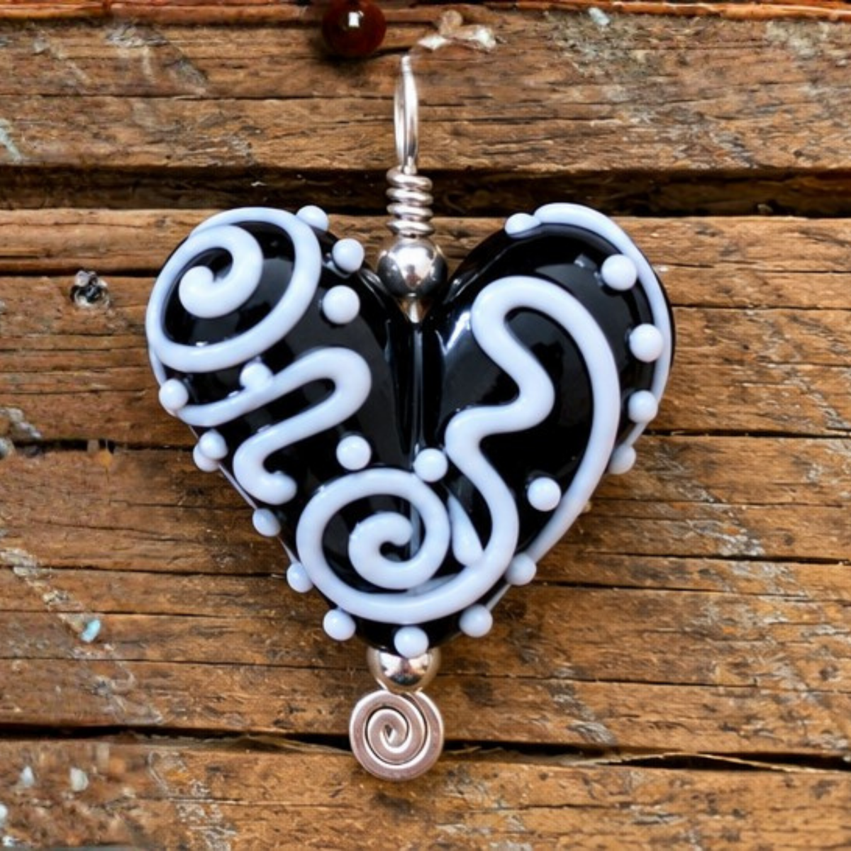 A heart-shaped glass pendant necklace with a black and white swirl pattern, featuring silver-toned metal wire-wrapping and a small spiral charm at the bottom, displayed on a rustic wooden background.