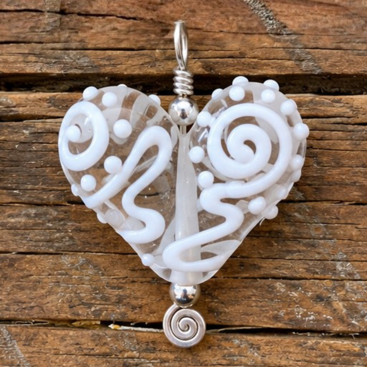A heart-shaped glass pendant with intricate white swirls and dots on a clear glass base, featuring silver-toned metal accents and a bail, displayed on a rustic wooden surface.