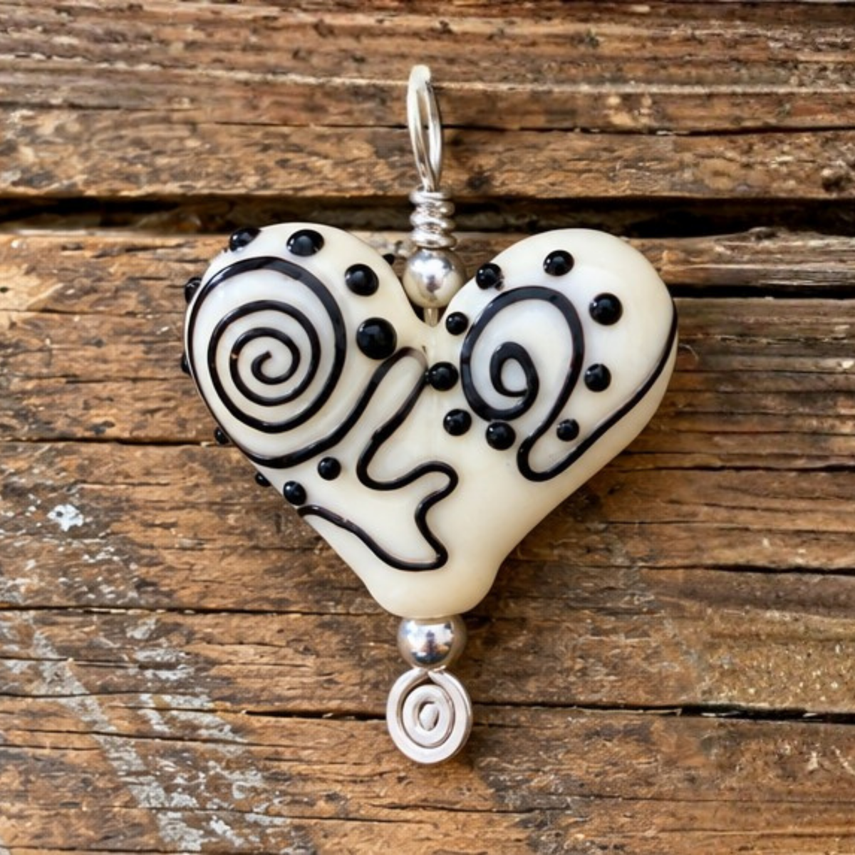 A heart-shaped glass pendant charm with black swirl and dot patterns on an off-white base, hanging from a silver bail over a rustic wooden background.