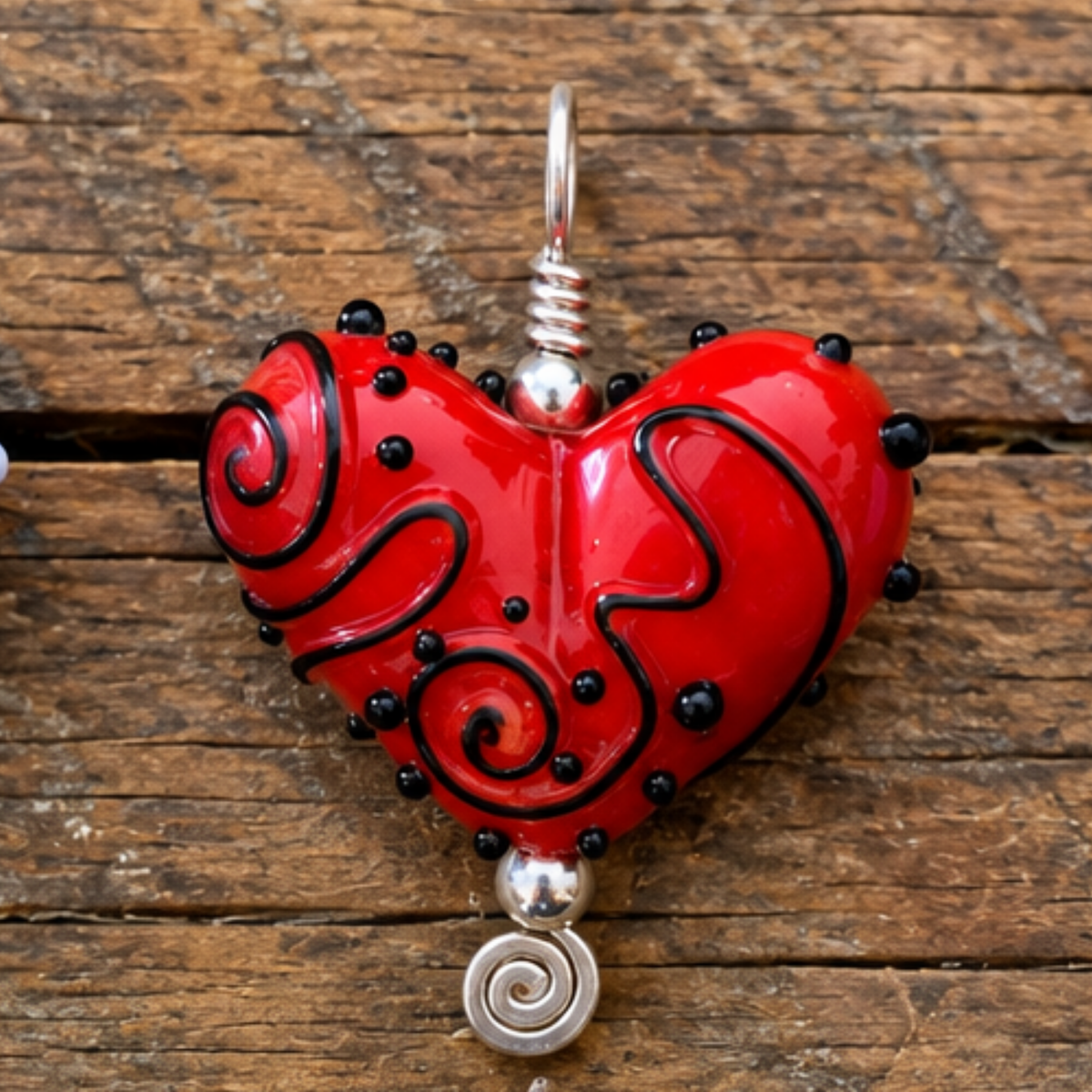 A red heart-shaped glass pendant with black swirls and small black beads, hanging from a silver metal loop against a rustic wooden background.