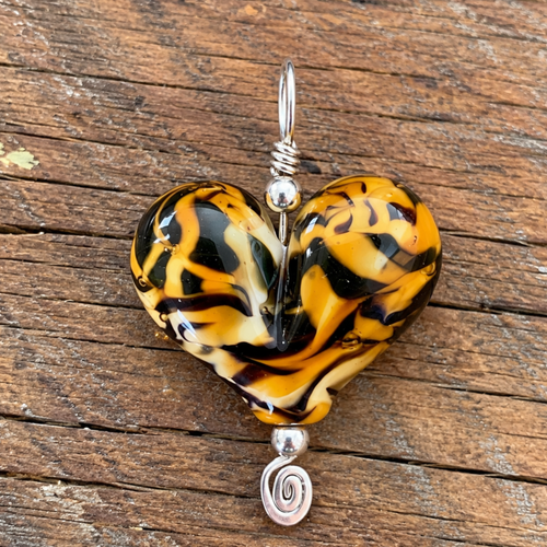 A heart-shaped glass pendant with a marbled black, orange, and yellow pattern, suspended from a silver wire loop with decorative beads, resting on a rustic wooden surface.