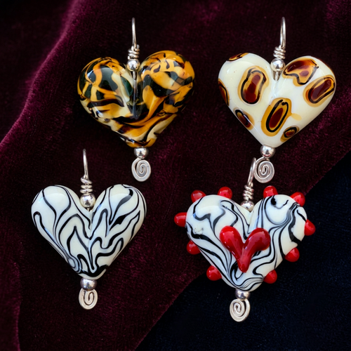 Four heart-shaped lampwork earrings displayed on a rustic wooden surface. The earrings feature various patterns and colors: one has a yellow, black, and white tiger-stripe pattern; one is cream with brown and black spots; one is cream with black wavy lines; and one is cream with black lines, red spikes, and a red heart in the center.