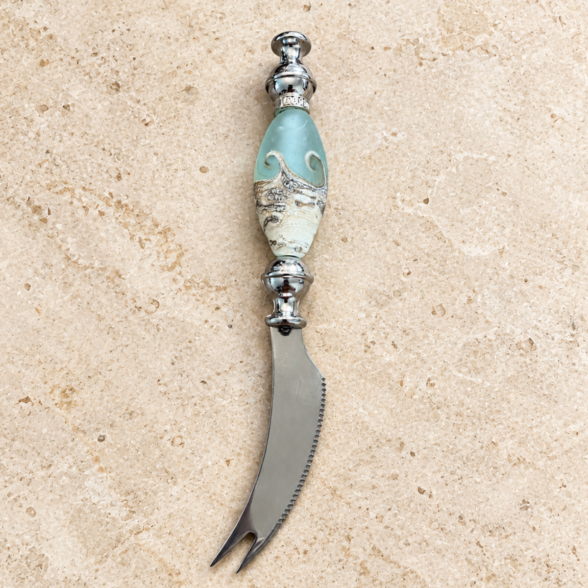 A decorative cheese knife with a curved silver steel blade and a handle featuring a light blue glass orb and a white marbled section, displayed on a beige stone surface.