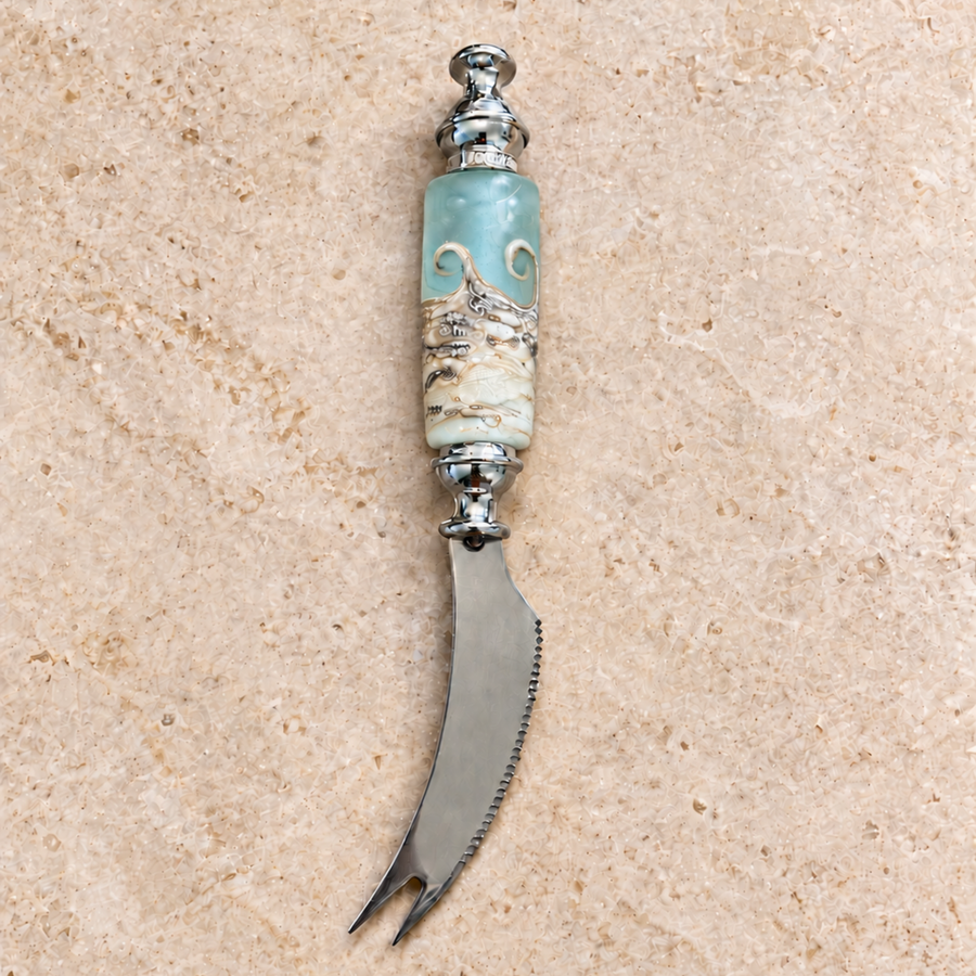 A decorative cheese knife with a curved silver steel blade and a carved handle featuring a light blue-green stone top and a cream-and-silver marbled lower section, displayed on a beige stone surface.