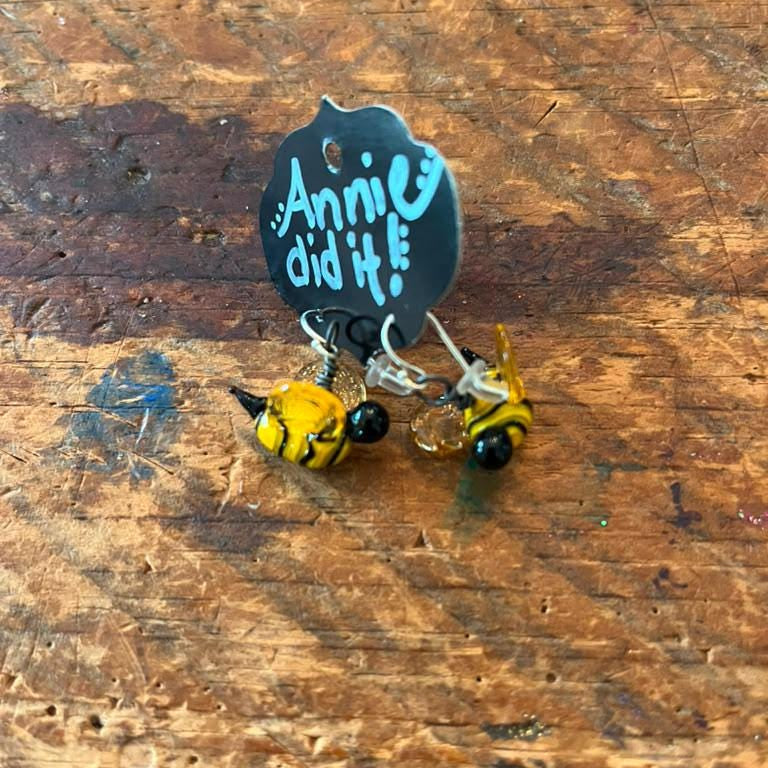 A pair of handmade bumblebee earrings with yellow and black bodies, displayed on a rustic wooden surface next to a black display card that reads 'Annie did it!'.