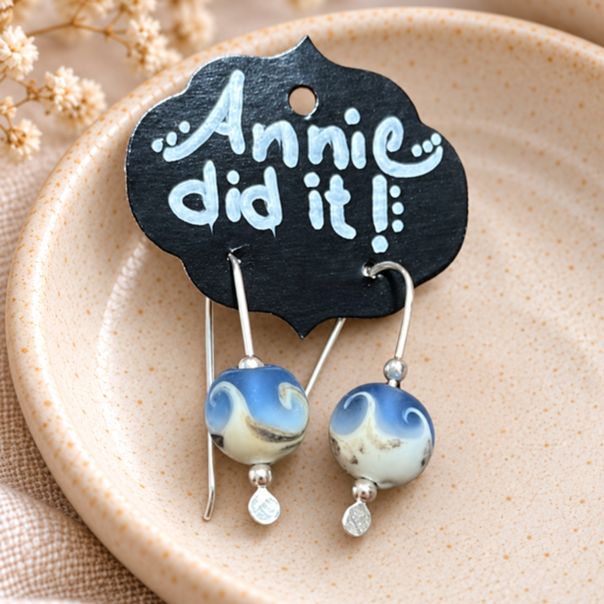 A pair of handmade dangle earrings with blue and white marbled glass beads and silver hooks, hanging from a black display card that reads 'Annie did it!'. The card is placed on a beige ceramic dish with dried flowers in the background.