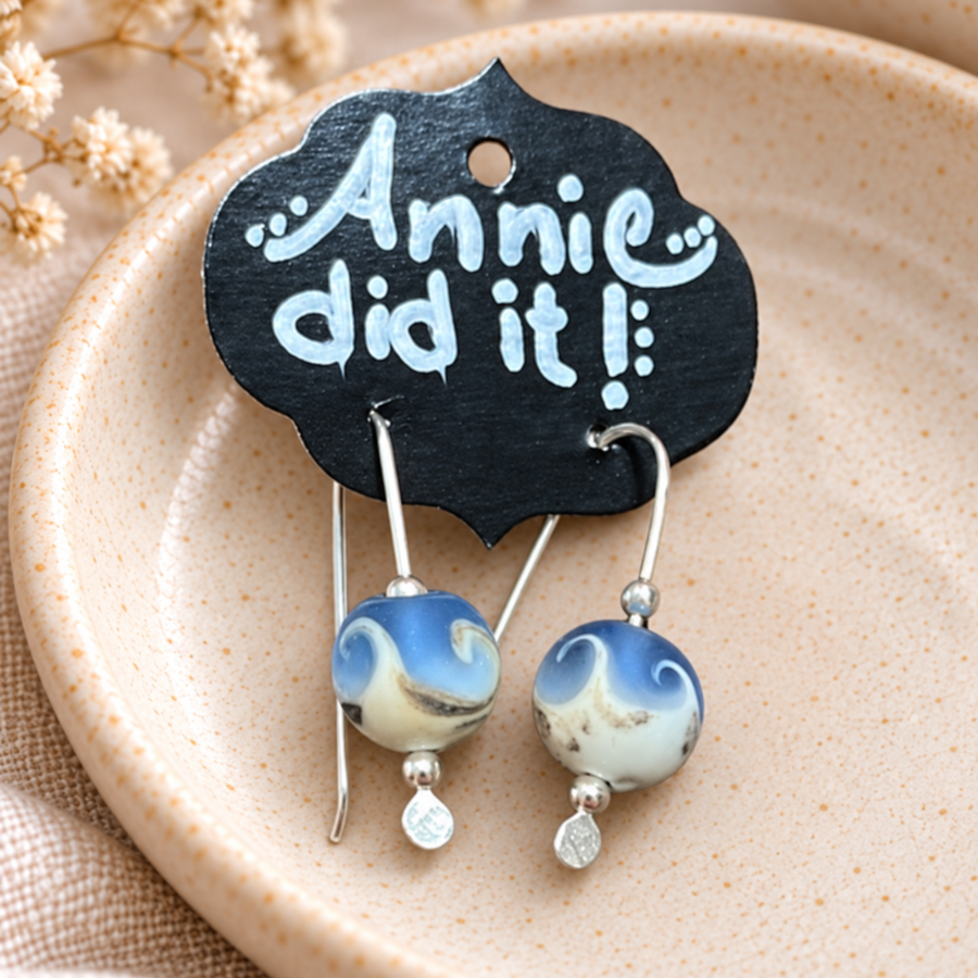 A pair of handmade dangle earrings with blue and white marbled glass beads and silver hooks, hanging from a black display card that reads 'Annie did it!'. The card is placed on a beige ceramic dish with dried flowers in the background.