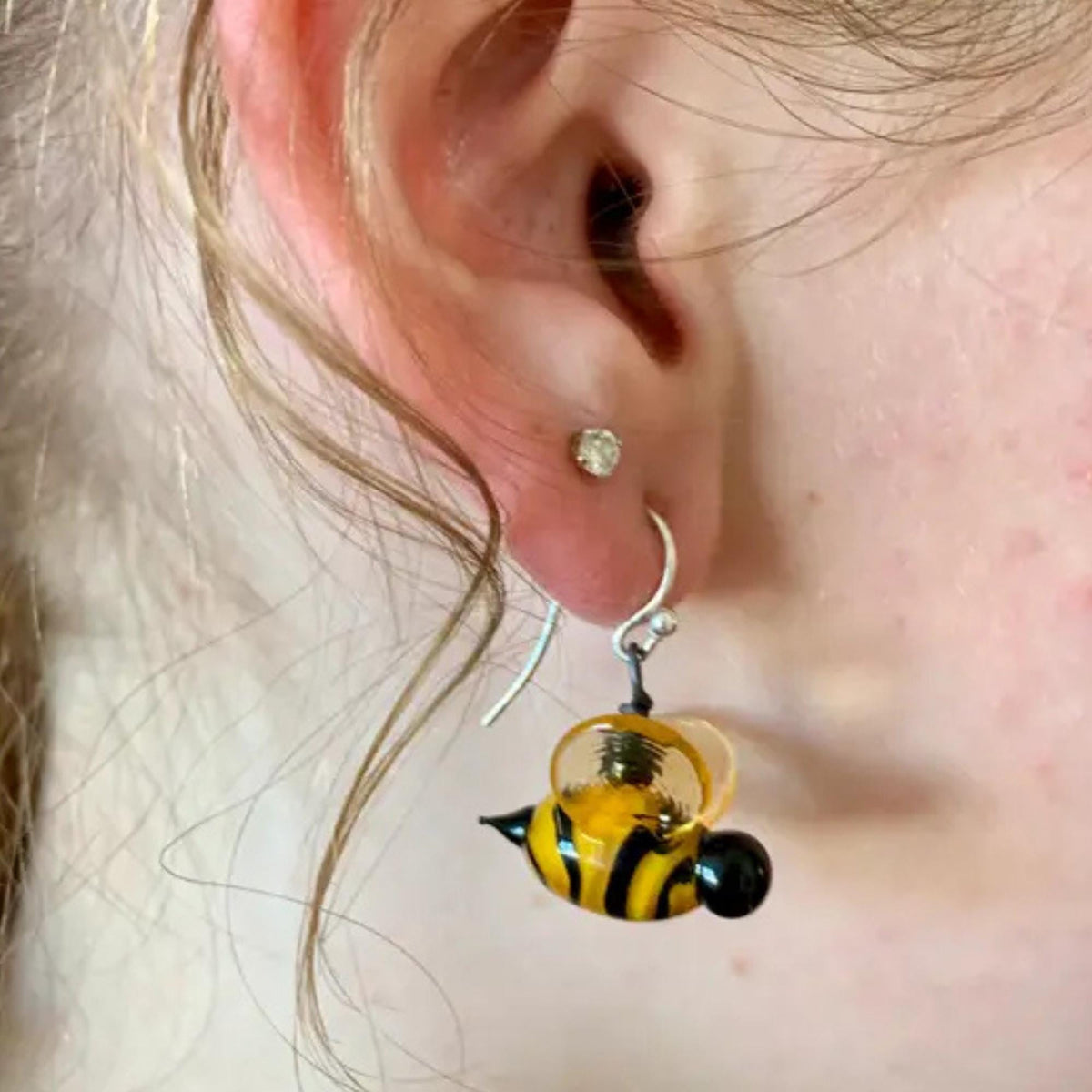 A close-up of a person's ear wearing a silver hoop earring with a dangling glass bee charm. The bee charm is yellow and black striped with a clear glass body and a small black bead tail.