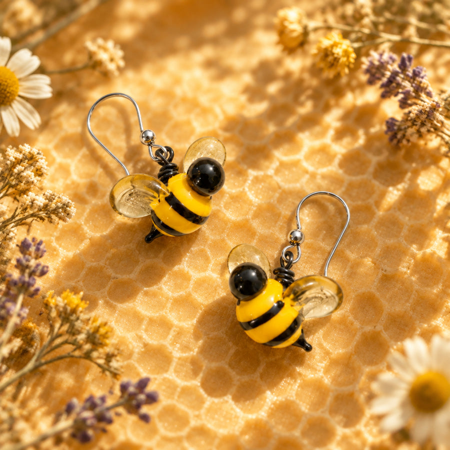 A pair of bumblebee-shaped glass dangle earrings with yellow and black bodies, clear wings, and silver fishhook wires, displayed on a textured off-white fabric background with soft leaf shadows. The brand logo 'Earrings' is visible in the top left corner.