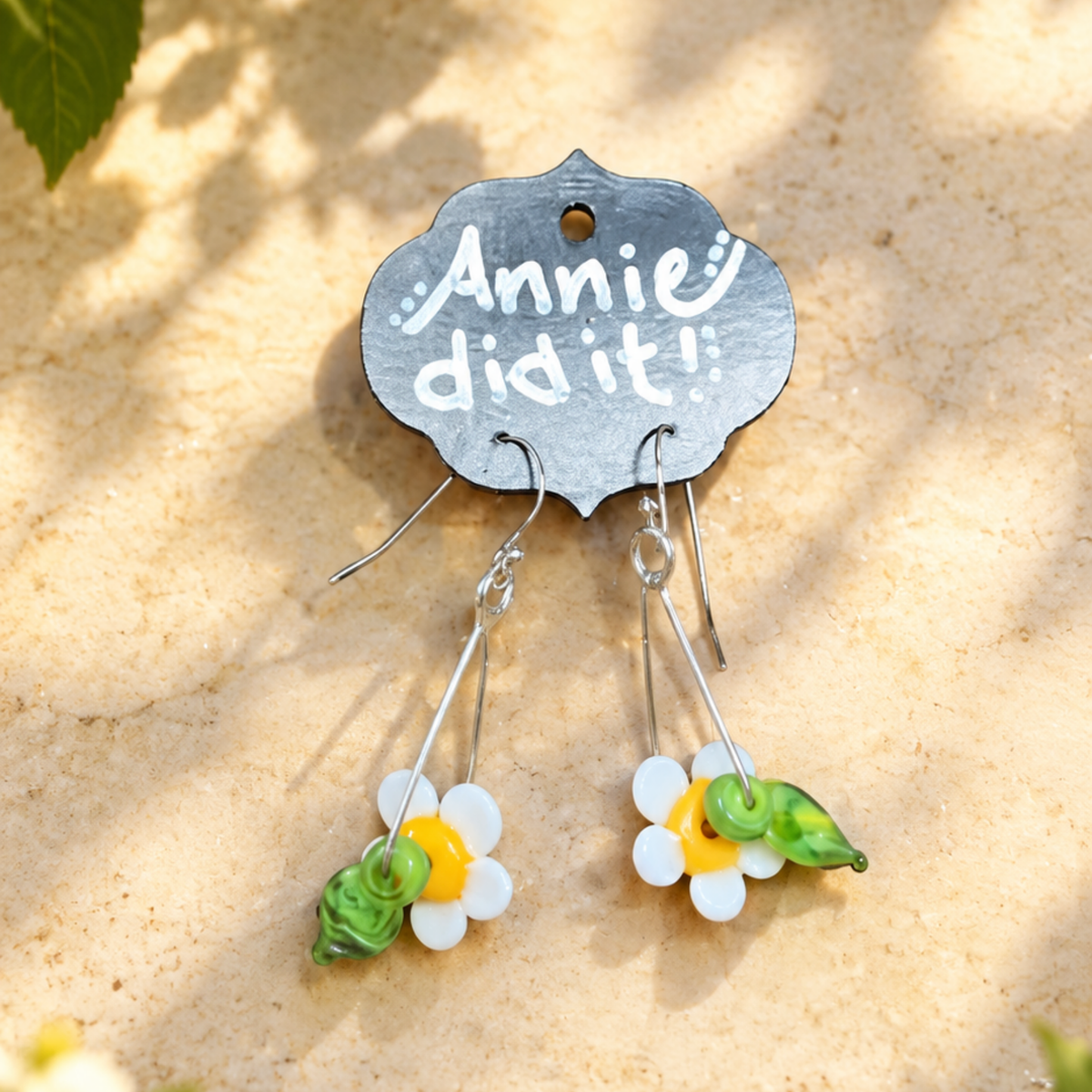 A pair of dangling earrings displayed on a silver card that reads 'Annie did it!'. The earrings have silver hooks and drop charms shaped like white daisies with yellow centers and green leaves, set against a sandy beige background with soft leaf shadows.