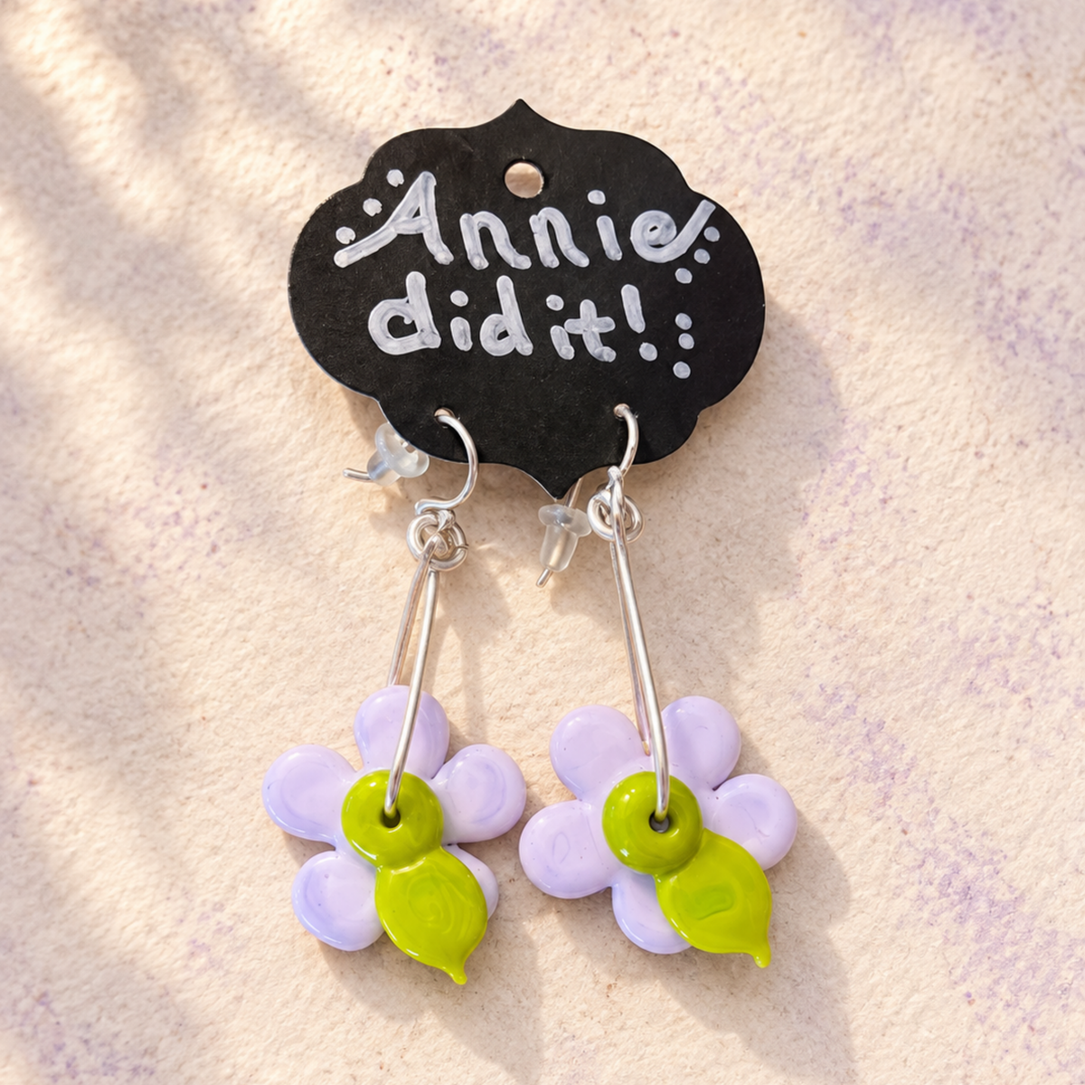 A pair of handmade dangle earrings featuring light purple flower charms with bright green centers, hanging from silver hooks and displayed on a black cloud-shaped card that reads 'Annie did it!'.
