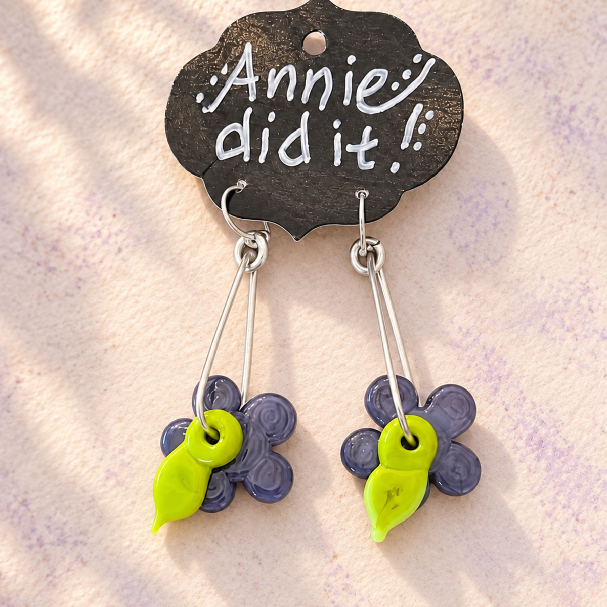 A pair of handmade dangle earrings with purple flower charms and bright lime green bead accents, hanging from silver-toned wires and displayed on a black decorative card that reads 'Annie did it!'.