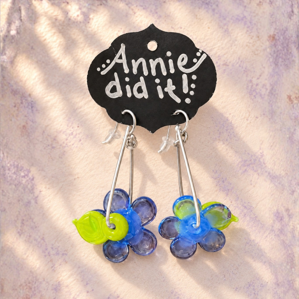 A pair of handmade dangle earrings displayed on a black card that reads 'Annie did it!'. The earrings feature blue and purple flower charms with bright green leaf accents and silver-tone hooks, set against a textured light background with soft leaf shadows.