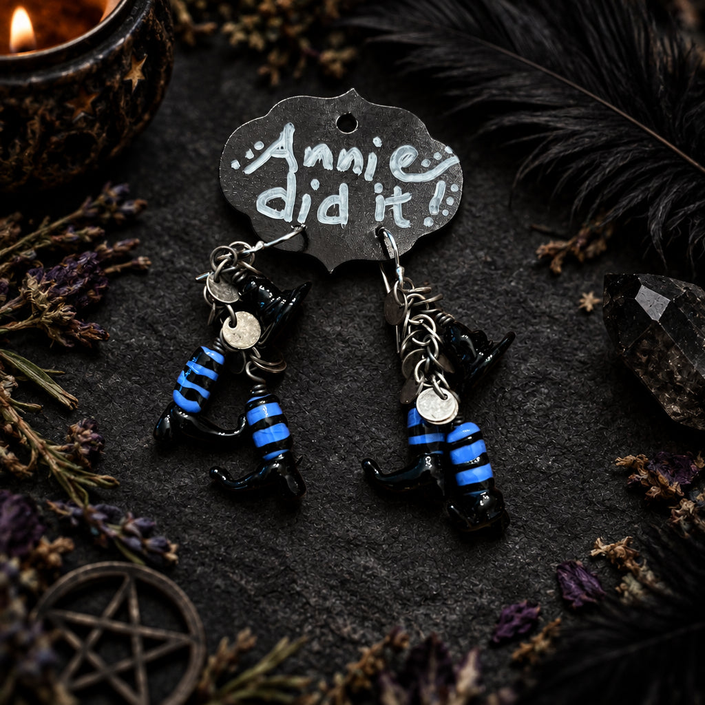 A pair of handmade dangle earrings featuring small black cat figurines wearing blue and black striped outfits, hanging from a silver-toned metal ring with a decorative tag that reads 'Annie did it!' on a rustic wooden surface.
