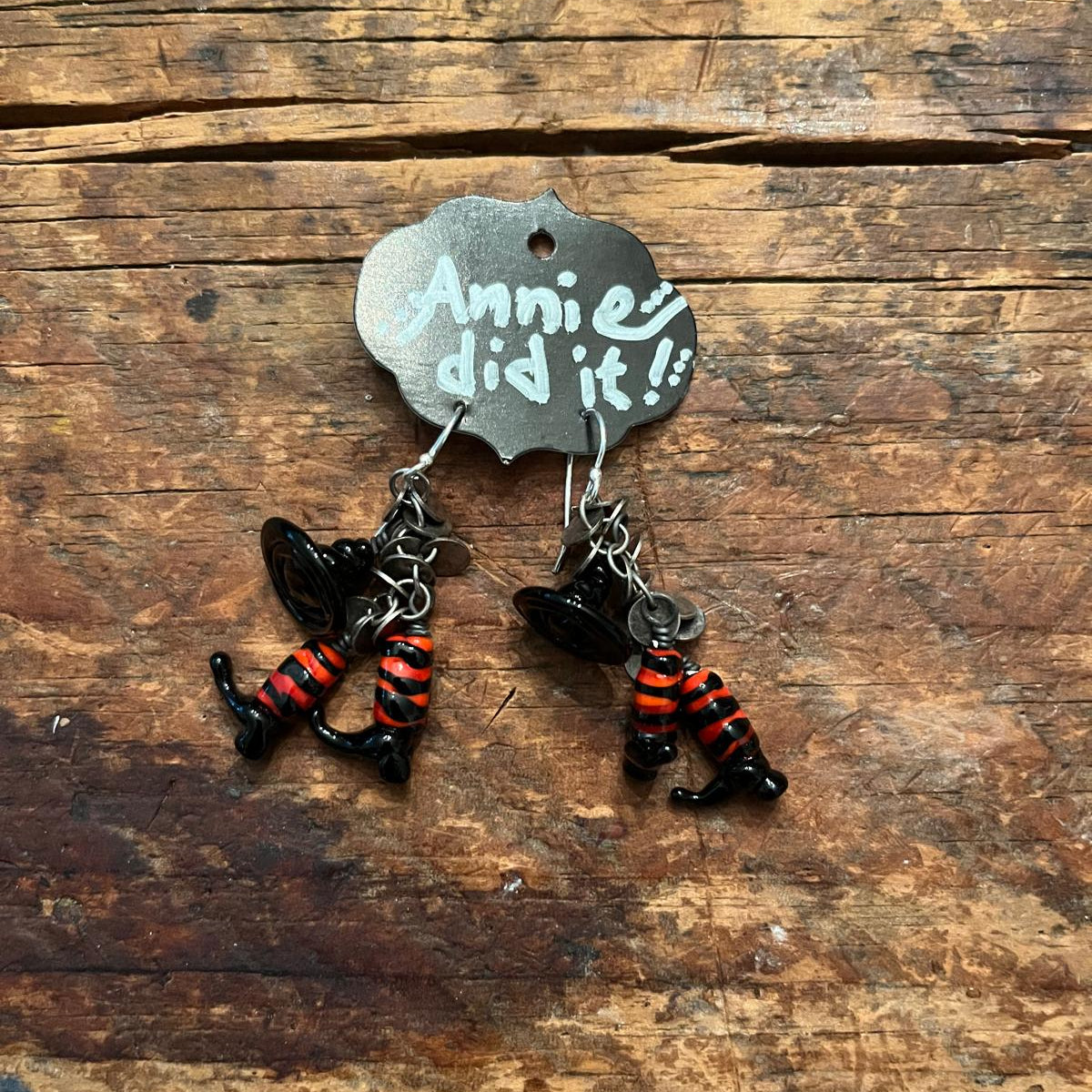 A pair of dangling earrings displayed on a small metal hanger that reads 'Annie's did it!!', placed on a rustic wooden surface. The earrings feature black witch hats and black-and-orange striped boot charms.