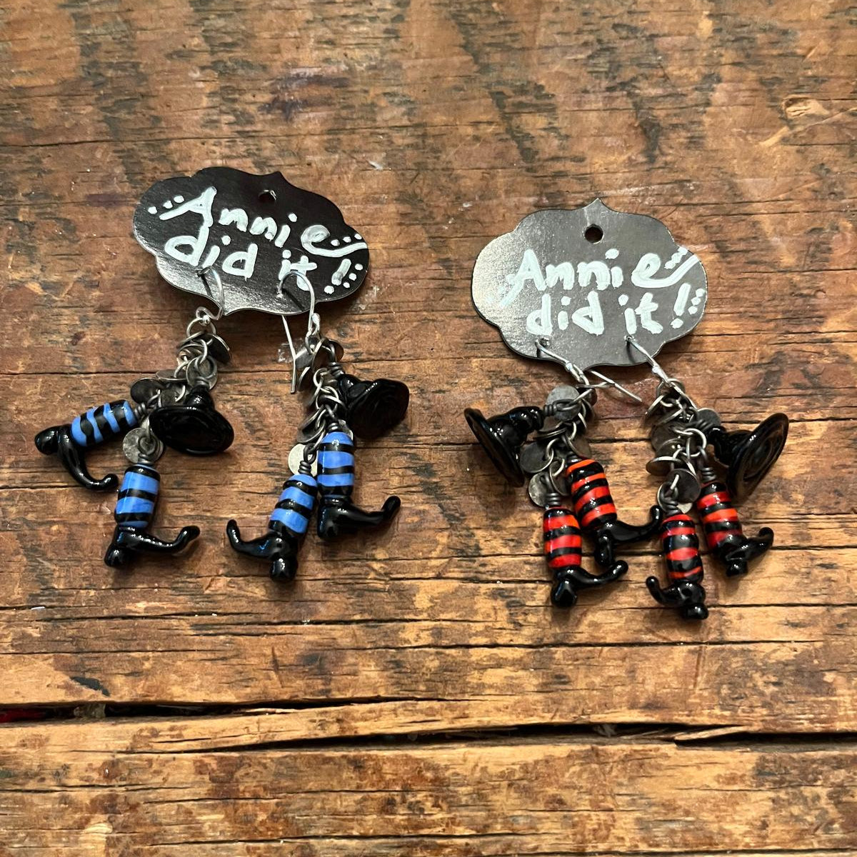 A pair of handmade dangle earrings featuring witch characters with striped pants, one pair with blue stripes and one pair with red stripes, hanging from silver hooks on a rustic wooden background. Each earring has a small silver cloud-shaped tag with the text 'Annie did it!!' written on it.
