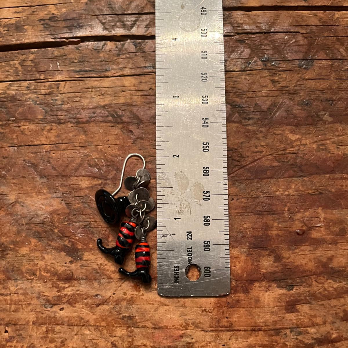 A small black and red striped fishing lure with a silver ear hook, shown next to a metal ruler on a rustic wooden surface.