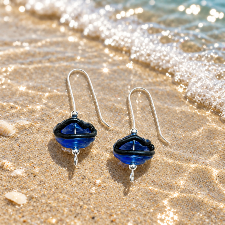 A pair of handmade glass drop earrings with deep blue and black swirls, suspended from silver fishhook wires, displayed on a textured off-white fabric background with soft leaf shadows.