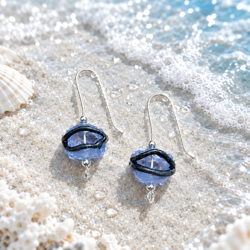 A pair of dangling earrings with silver-tone fishhook wires and oval blue beads outlined in black, shown on a light textured fabric background with soft leaf shadows. The text 'Earrings Splashy Night Blue' appears in the upper left.