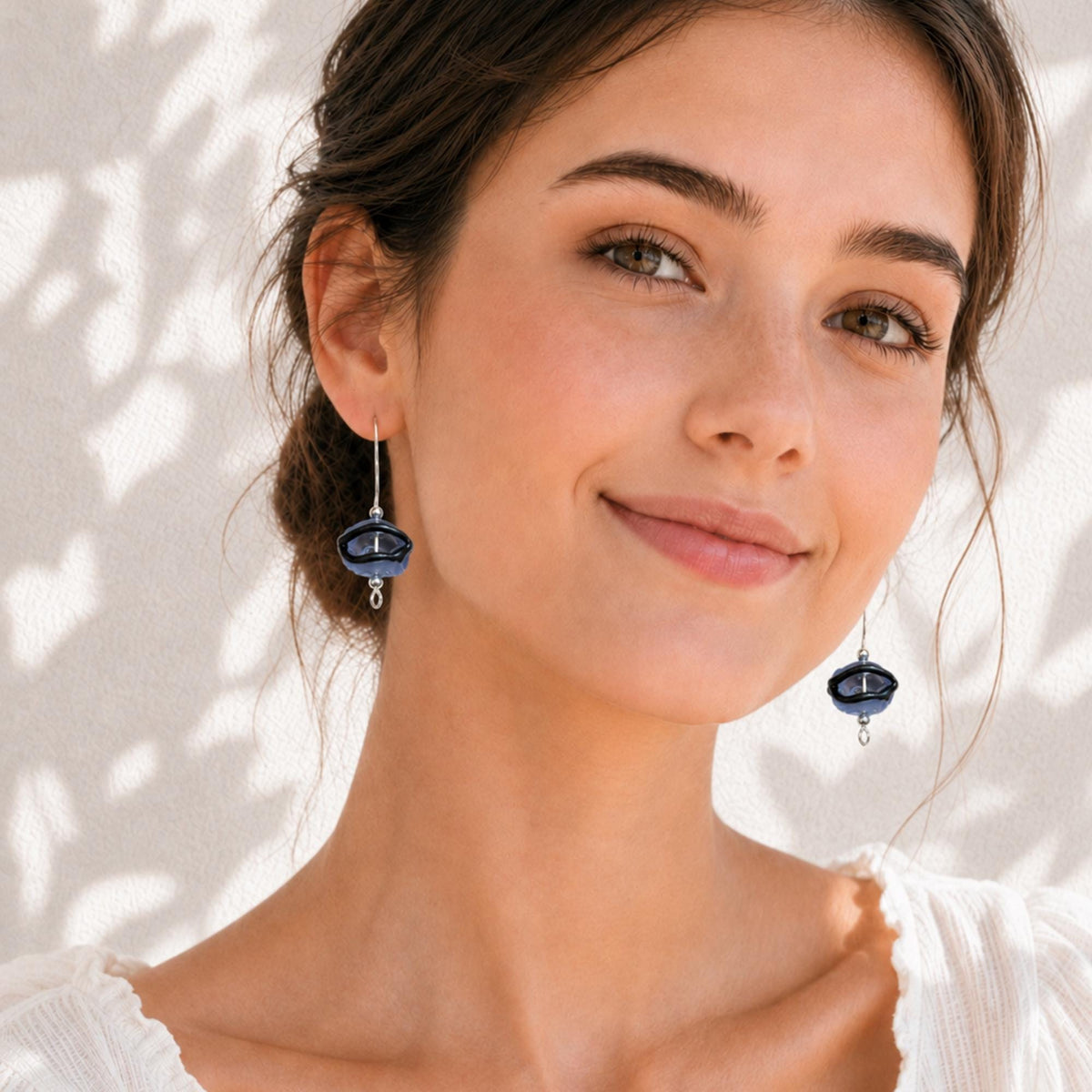A close-up lifestyle shot of a woman wearing matching dark blue and silver dangle earrings, set against a warm off-white wall with soft leaf shadows.