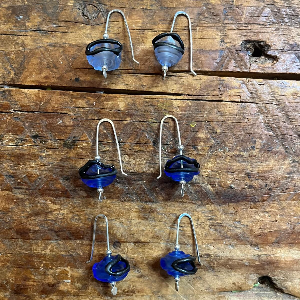 Six handmade glass bead drop earrings with silver-toned fishhook wires, featuring blue and black swirled beads, arranged on a rustic wooden surface.