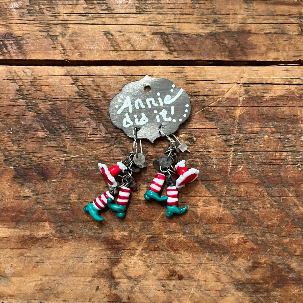 A pair of handmade dangle earrings displayed on a metal sign that reads 'Annie did it.' The earrings feature red and white striped socks with teal boots, shown on a rustic wooden surface.