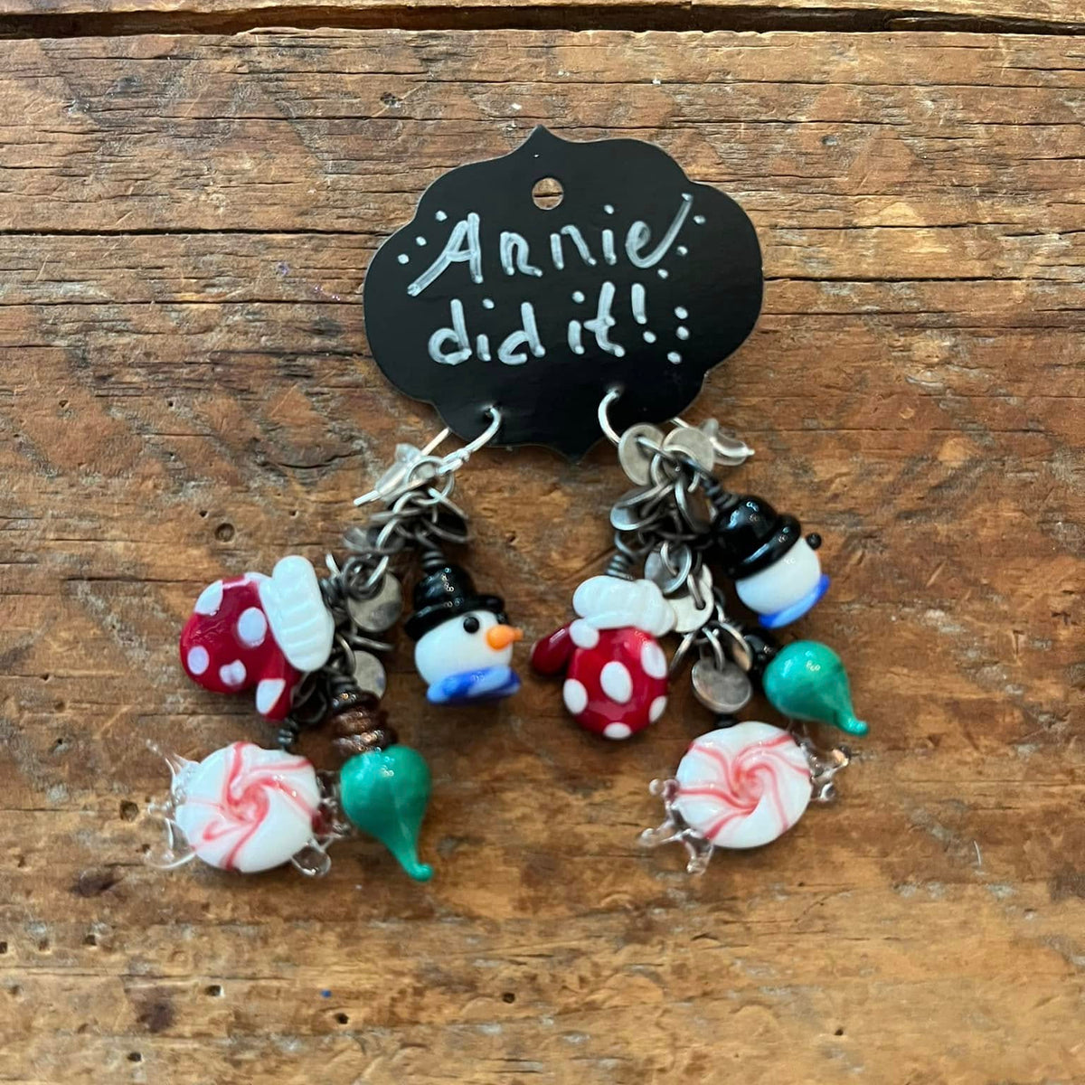 A pair of handmade Christmas-themed dangle earrings displayed on a small black chalkboard sign that reads 'Annie did it!'. The earrings feature silver-toned chains with charms including red-and-white peppermint candies, green and teal Kiss Kiss candies, snowman figures, and candy canes, resting on a bed of faux snow with blurred holiday lights and evergreen branches in the background.