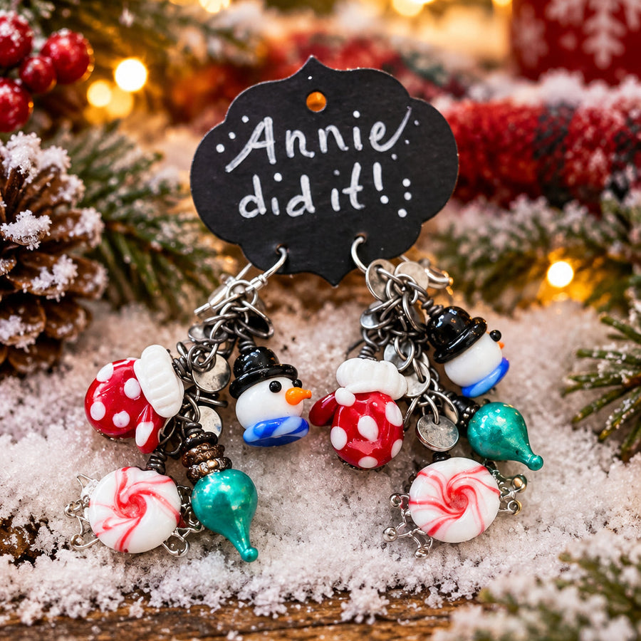 A pair of handmade Christmas-themed dangle earrings displayed on a small black chalkboard sign that reads 'Annie did it!'. The earrings feature silver-toned chains with charms including red-and-white peppermint candies, green and teal Kiss Kiss candies, snowman figures, and candy canes, resting on a bed of faux snow with blurred holiday lights and evergreen branches in the background.
