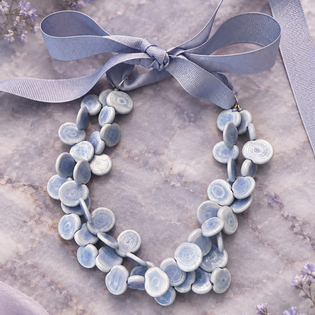 A handmade statement necklace made of light blue and white ceramic flower petals, tied with a wide lavender ribbon bow, displayed on a rustic wooden surface.
