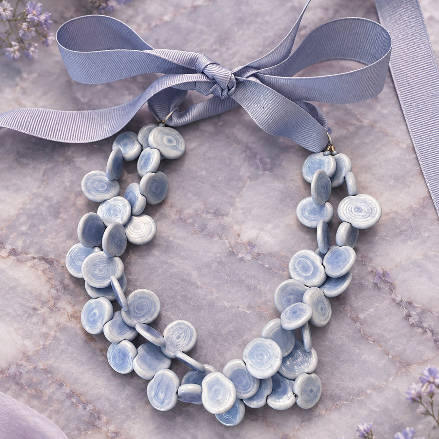 A handmade statement necklace made of light blue and white ceramic flower petals, tied with a wide lavender ribbon bow, displayed on a rustic wooden surface.