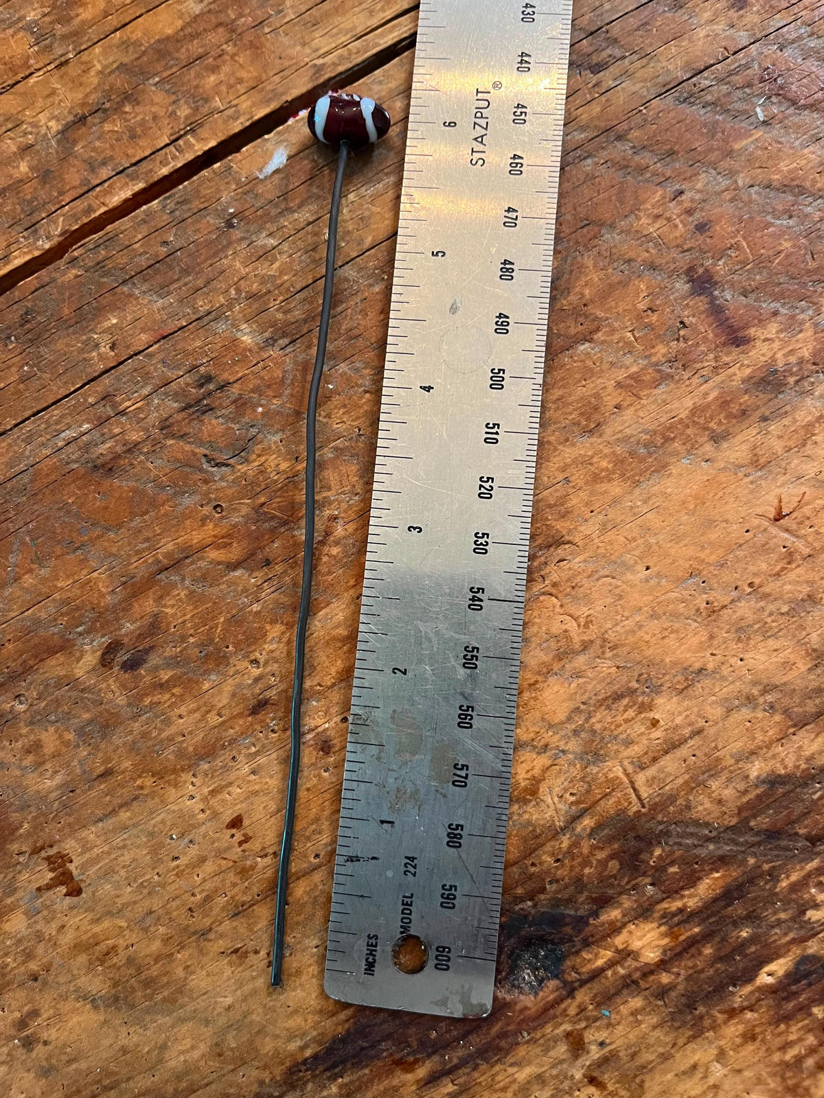 A small cylindrical electronic component with a long thin lead lies next to a metal ruler on a rustic wooden surface.
