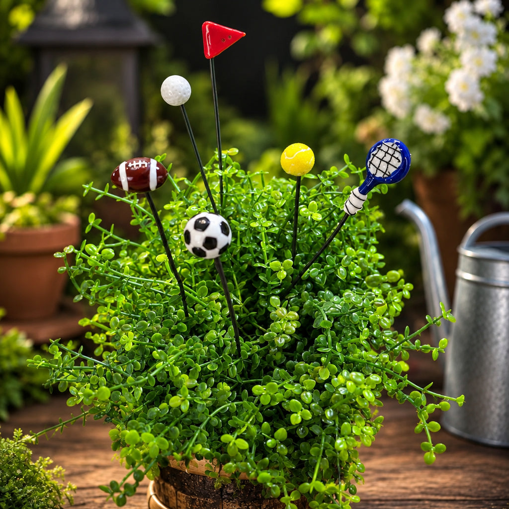 A set of sports-themed plant stakes with small decorative balls and miniature sports objects, displayed in a green succulent plant. The set includes a football, soccer ball, tennis racket, tennis ball, and flag on a wooden table with a watering can and potted plants in the background.