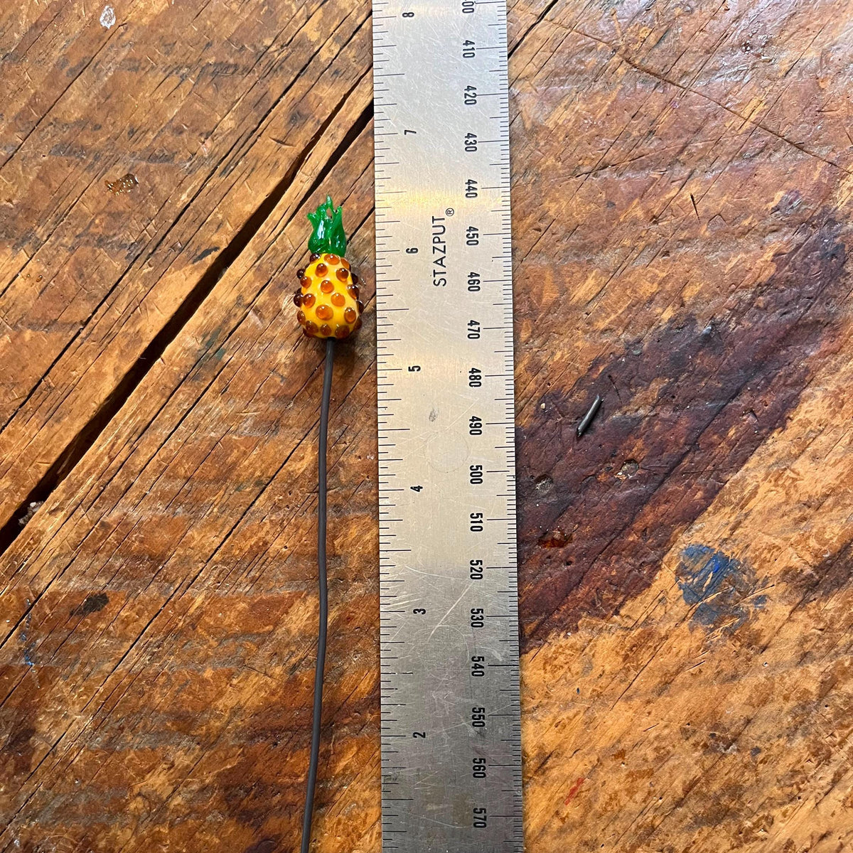 A small handmade bead accessory with a yellow and orange pineapple-shaped charm on a long black wire, shown next to a metal ruler on a rustic wooden surface.