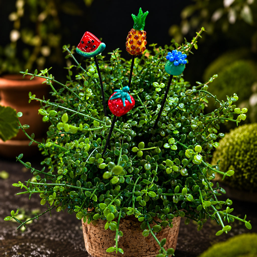 A set of five colorful plastic plant stakes shaped like a watermelon, pineapple, blue flower, red strawberry, and kiwi, shown inserted into a small potted green plant. The brand logo 'Plant Sticks Fruit Bundle' is visible in the top left corner.