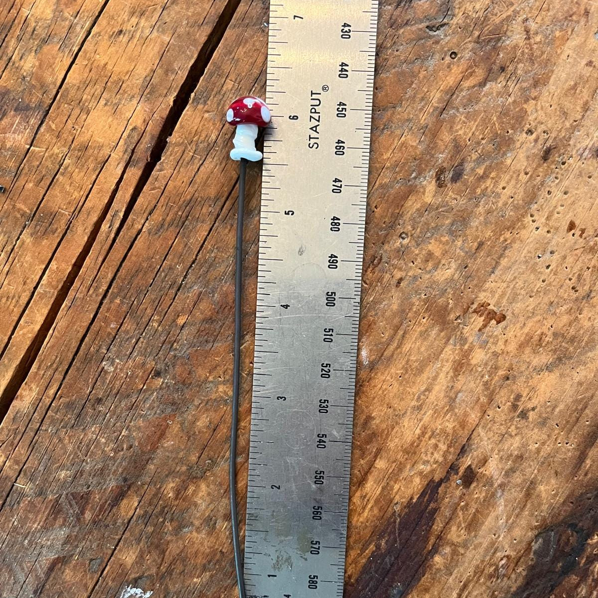 A small red and white mushroom-shaped bead or charm on a long black cord, shown next to a metal ruler on a rustic wooden surface.