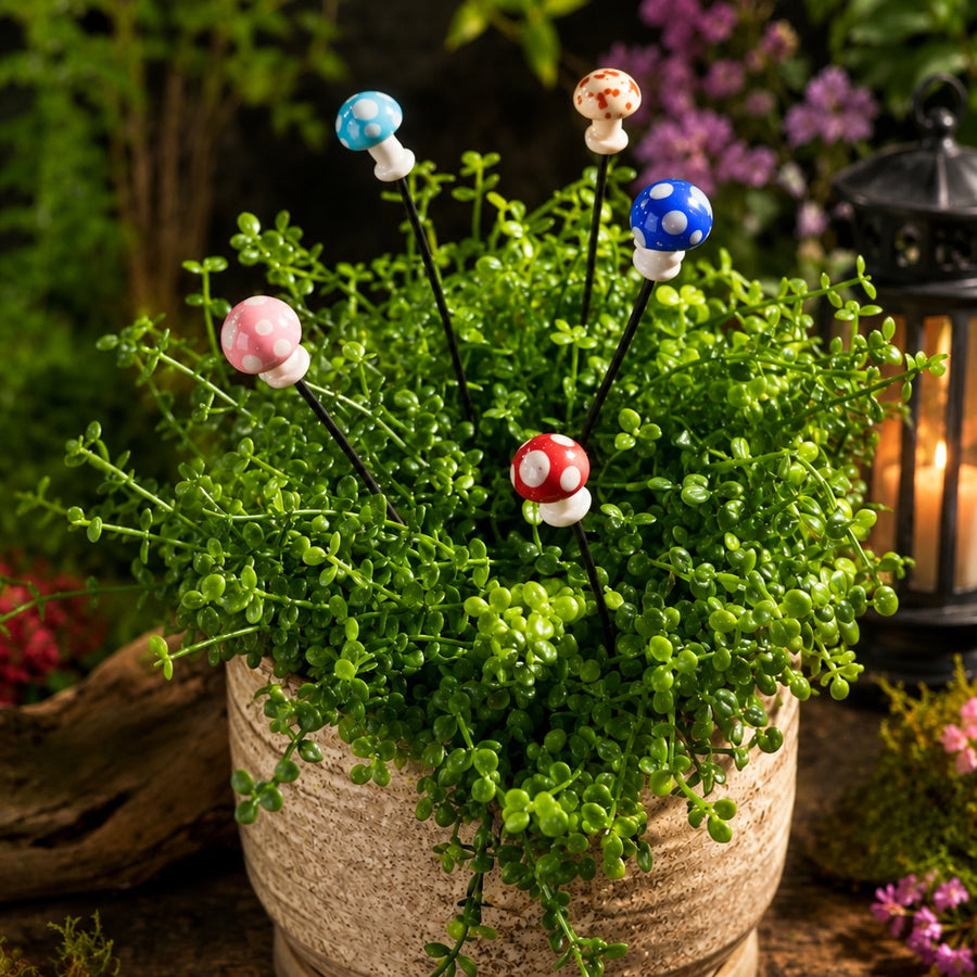 Five mushroom-shaped plant sticks with colorful ceramic caps are shown planted in a small green succulent arrangement in a beige pot, with a blurred garden background and a lantern. The logo 'Plant Sticks Mushroom Bundle' appears in the top left.