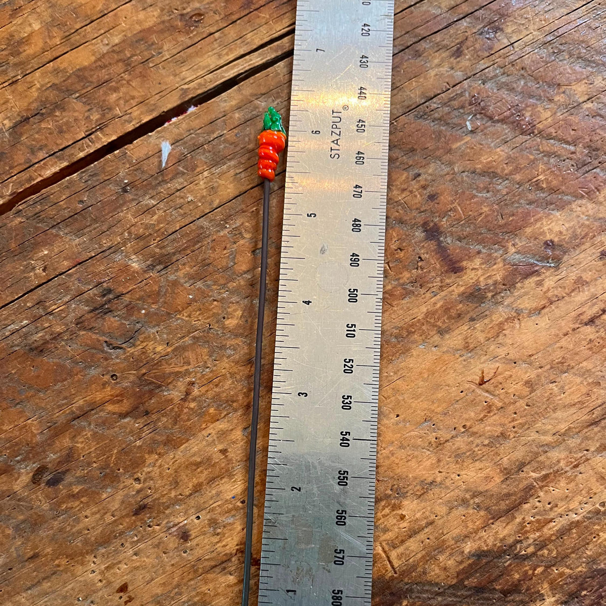 A handmade beaded pin with a long dark metal shaft and a small red and green beaded top, shown next to a metal ruler on a rustic wooden surface.