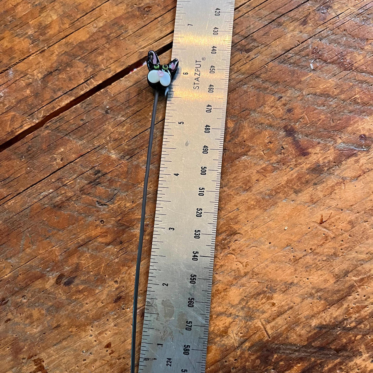 A small handmade jewelry piece with a long black wire and two light blue beads, shown next to a metal ruler on a rustic wooden surface.