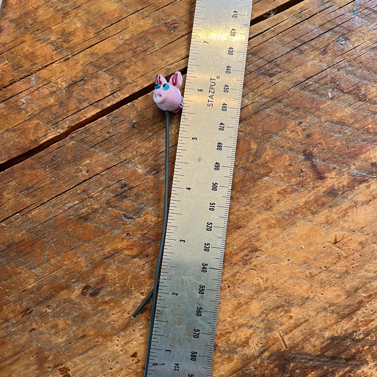 A small pink bunny-shaped bead or charm on a long black string, shown next to a metal ruler on a rustic wooden surface.