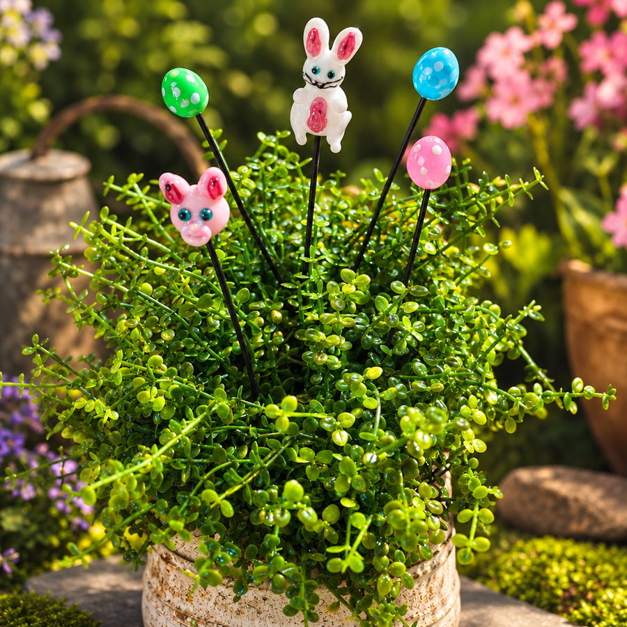 Easter plant stake bundle with decorative garden sticks featuring a white bunny, pink pig, green egg, and blue egg on black stakes, shown inserted into a small potted green plant in a garden setting. The text 'Plant Sticks Easter Bundle' appears in the top left.