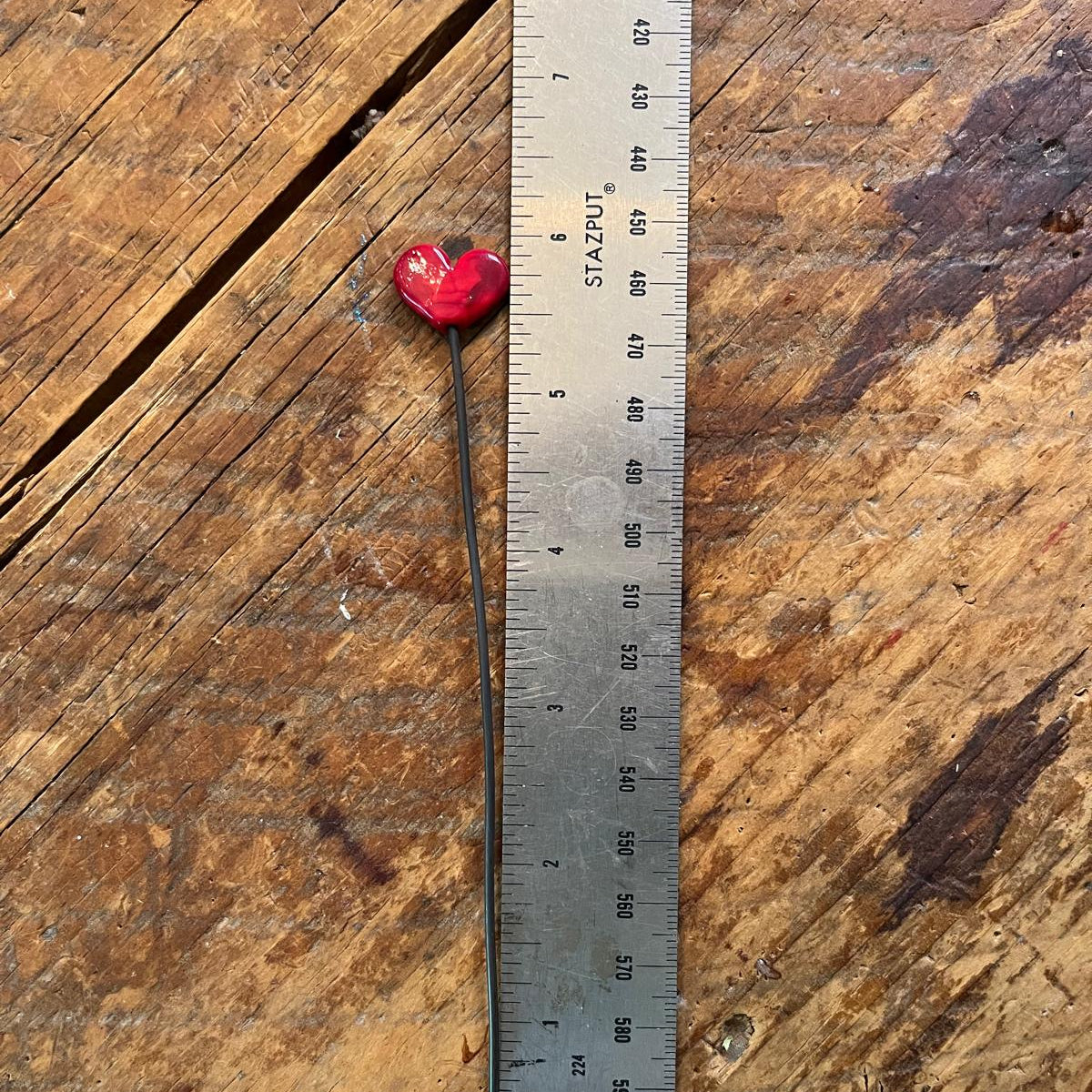 A small red and black fishing lure or hook shown next to a metal ruler on a rustic wooden surface.