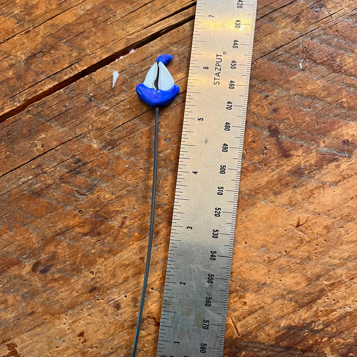 A small blue and white decorative pin with a long dark metal stem lies on a rustic wooden surface next to a metal ruler for scale.