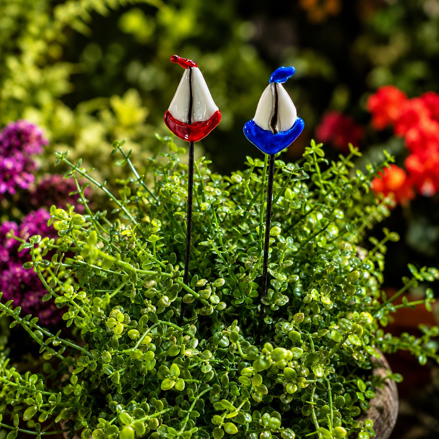 Two decorative garden soil stakes shaped like small boats, one red and white and one blue and white, planted in a green leafy plant arrangement. The Plant Sticks logo is visible in the top left corner.