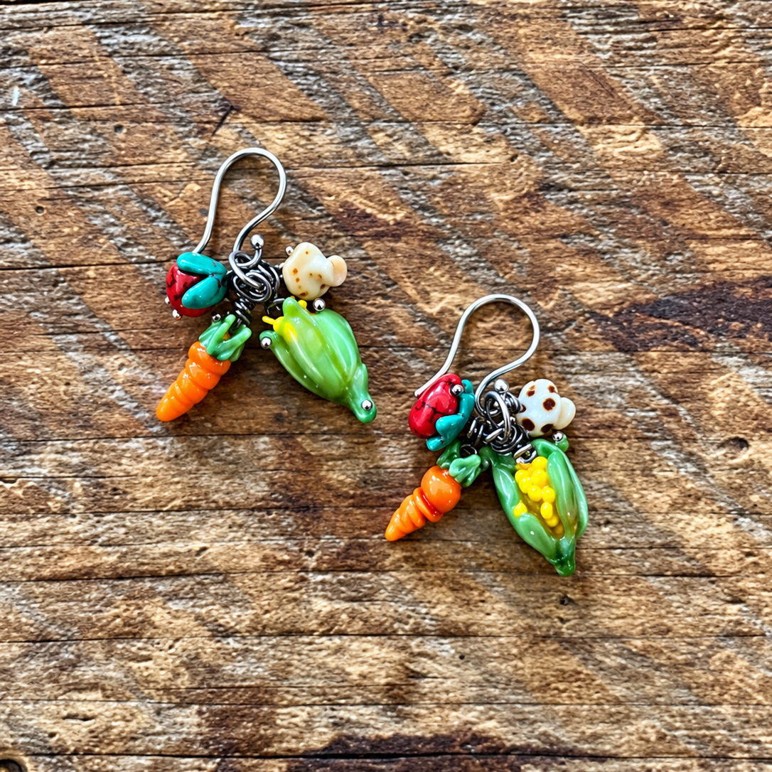 A pair of handmade vegetable-themed dangle earrings resting on a rustic wooden surface. The earrings feature silver-toned fishhook wires with dangling charms shaped like carrots, peas, corn, and tomatoes in bright colors.