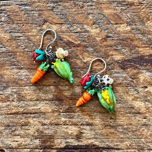 A pair of handmade vegetable-themed dangle earrings resting on a rustic wooden surface. The earrings feature silver-toned fishhook wires with dangling charms shaped like carrots, peas, corn, and tomatoes in bright colors.