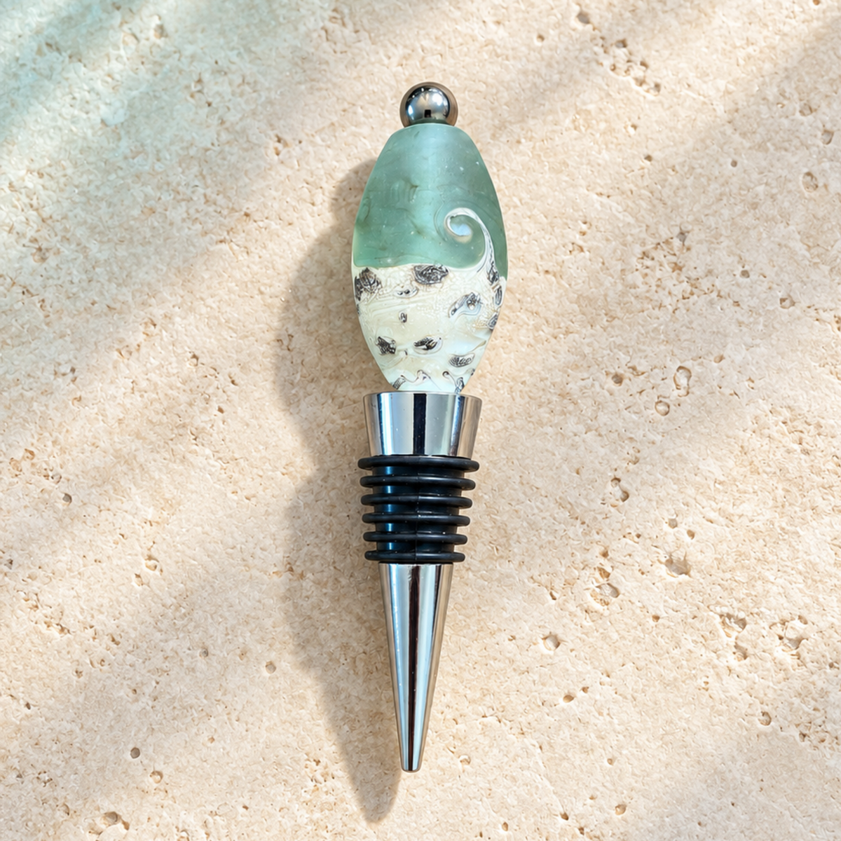 A handmade glass bottle stopper featuring a natural sea urchin shell bead with green and white patterns, a silver metal ball finial, and a black and silver rubber-and-metal bottle stopper base, set against a textured beige stone background.