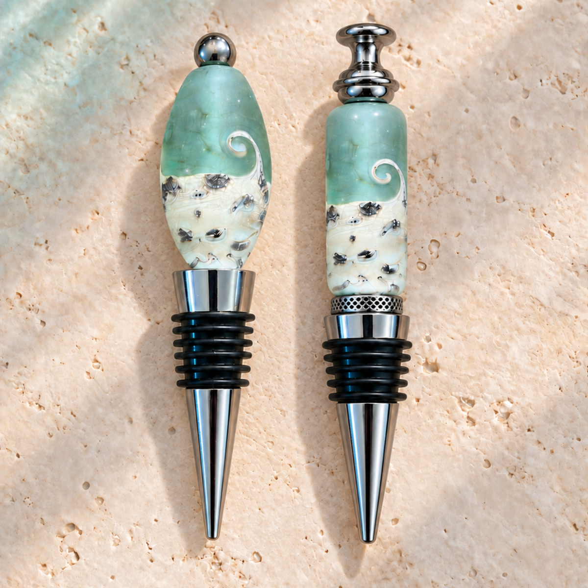 Two handmade glass wine stoppers with light green and white wave-patterned tops and silver metal collars, displayed on a textured beige stone surface.